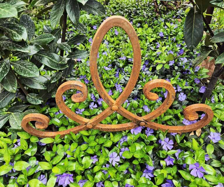 Szépen megmunkált kovácsoltvas dísz a zöld növények és lila virágok között.